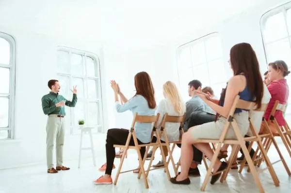 New Work e.V. – Moderner Workshop in einem lichtdurchfluteten Raum. Ein Redner in grüner Hemdbluse steht vor einer Gruppe von Zuhörern, die auf Holzstühlen sitzen und applaudieren. Die Atmosphäre ist offen und inspirierend.